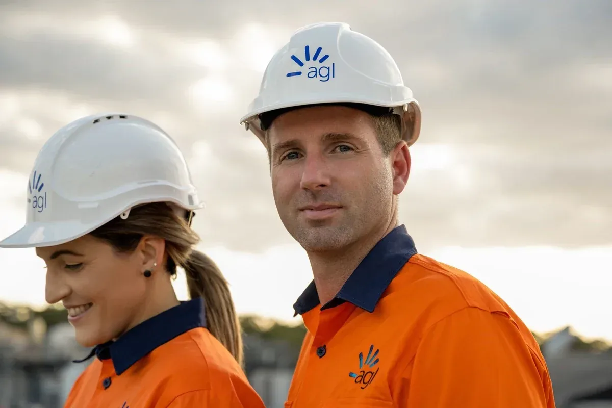 Two AGL workers in hi-vis shirts and branded hard hats on site under a cloudy sky